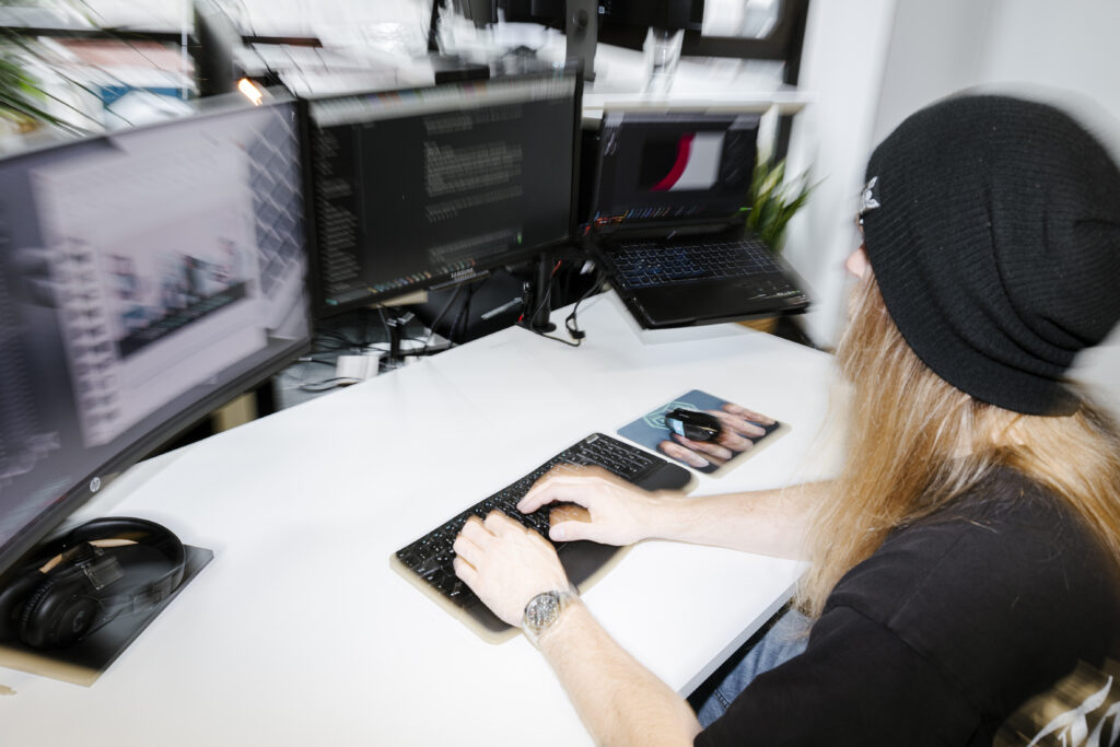 Schreibtisch mit Bildschirmen und Laptop und ein Mensch der mit einem Stuhl dran sitzt